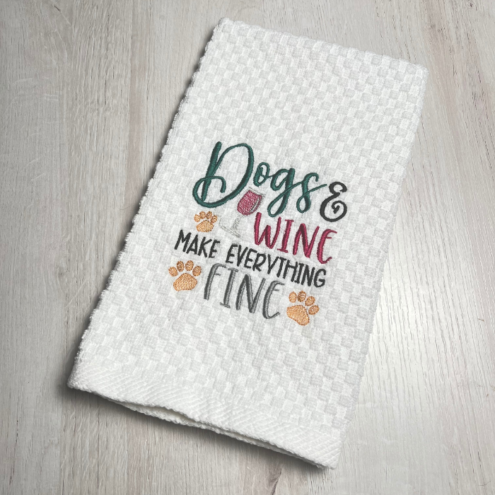 Embroidered white hand towel with saying "Dogs and wine make everything fine," glass of wine and 3 pawprints.