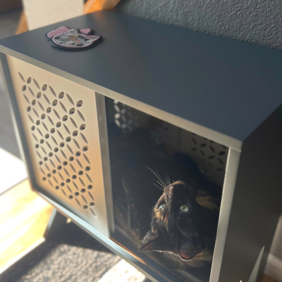 Grey table with white sliding doors and a cat bed shelf.  Black and tan tortie cat hanging out of the cube. Black and pink cat head shaped coasters lay on top of table.