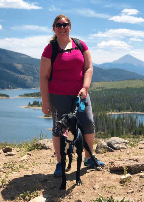 Woman exploring the world with her dog. She is in a pink shirt and capris with her black and white dog on the top of a hill in the mountains, overlooking a lake and forest.