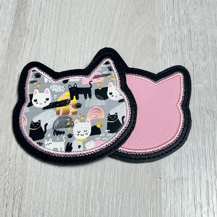 Two vinyl coasters in the shape of a cat head. The first coaster has a black, grey, white and pink cat collage vinyl stitched with pink thread onto a black vinyl backing. The second coaster lies underneath and is light pink stitched with pink thread to a black backing.