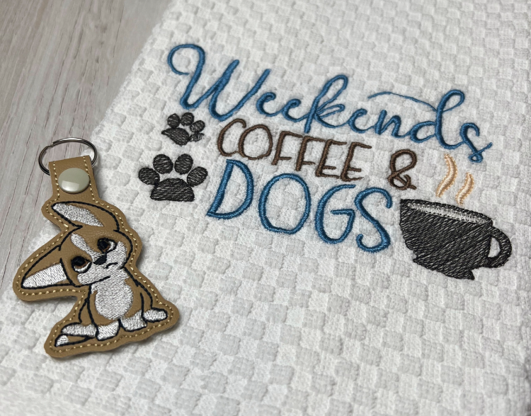 White embroidered towel with saying "Weekends, Coffee & Dogs" in blue and brown colors. Two brown pawprints on the left, and a brown cup of coffee with steam on the right. An embroidered, tan marine vinyl keychain of a dog with one ear up listening lays on top of the towel.